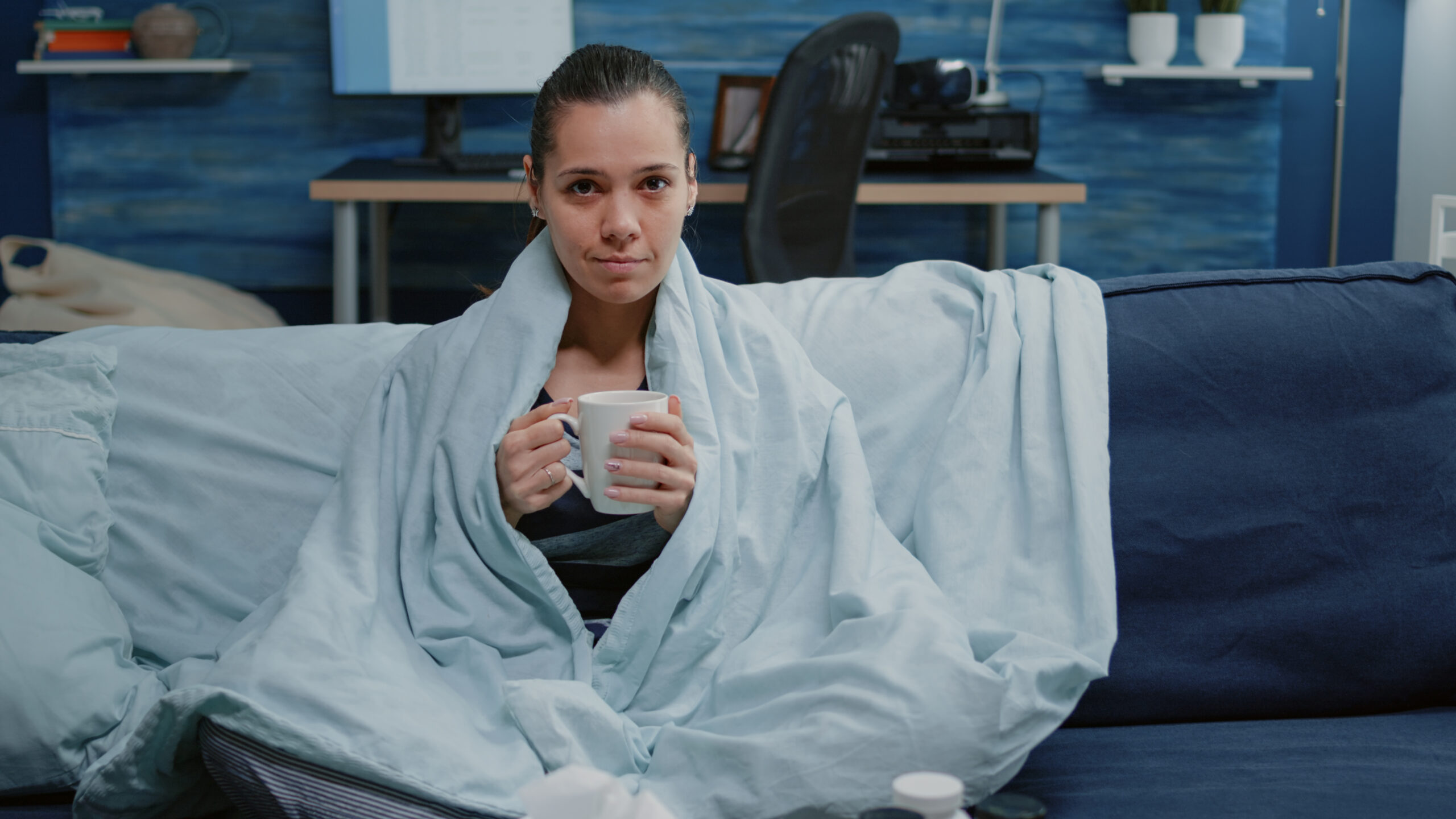 A woman sits on a couch wrapped in a light blue blanket, holding a white mug—perhaps taking an otc mucolytic as she copes with phlegm and cough. She looks at the camera with a neutral expression; a desk and computer are visible in the background.