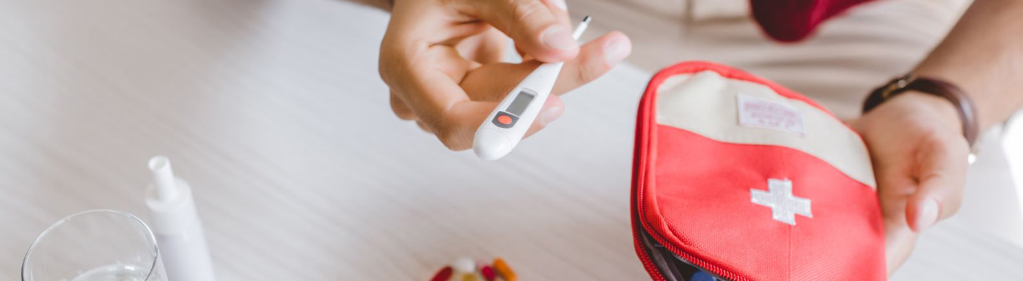 A person holds a digital thermometer and a red first aid kit with a white cross, while otc mucolytic medicine and a glass of water for chest congestion sit on the table nearby.