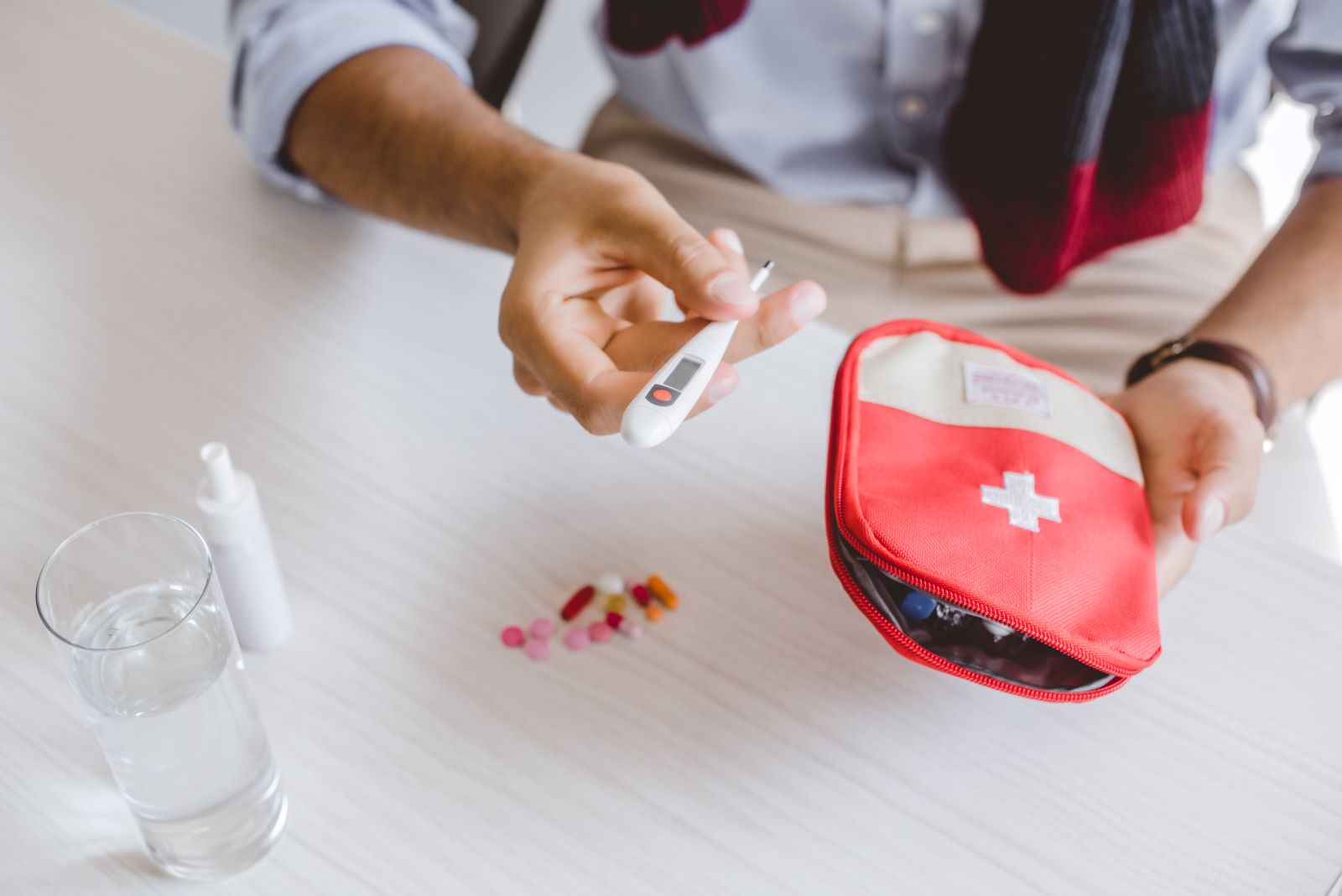 A person holds a thermometer and a red first aid kit on a table, next to a glass of water, some pills, phlegm medication, and a nasal spray.