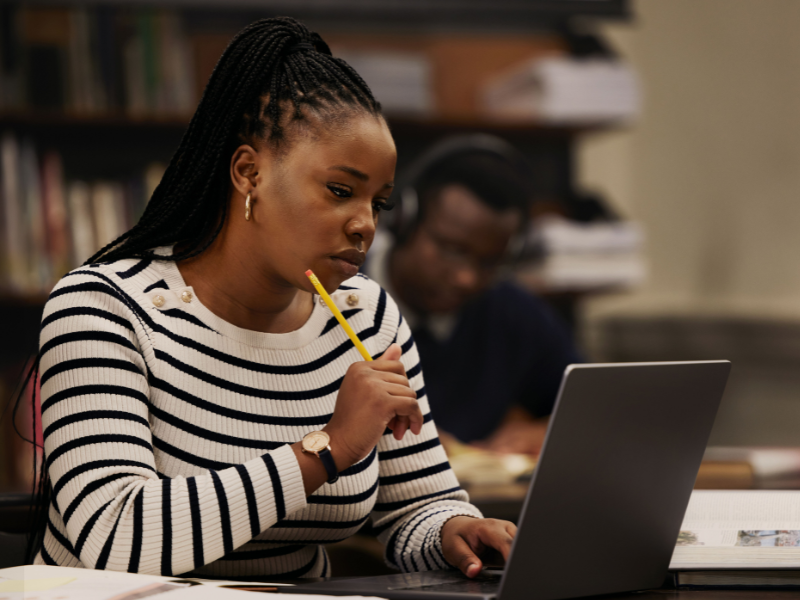 A woman has avoided the flu and can write her exam. She is wearing a striped sweater sits at a desk, looking thoughtfully at a laptop while holding a pencil to her chin. There are books and shelves in the background.