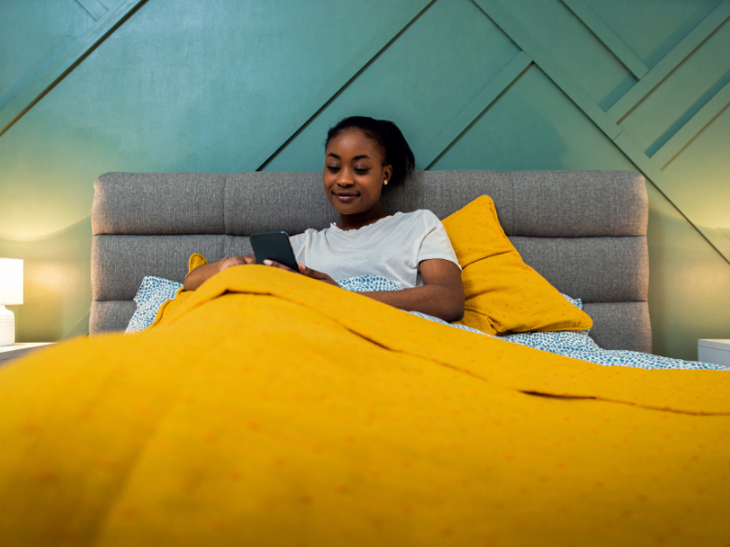 A woman avoiding the flu, sits up in bed under a yellow blanket, smiling while looking at her smartphone. The bed has a gray headboard and the room features modern, geometric wall decor.