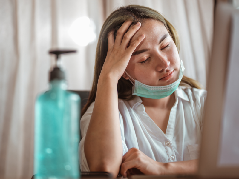 A woman wearing a face mask sits with her eyes closed, resting her head on her hand, thinking about how to avoid the flu. A bottle of hand sanitizer is visible in the foreground.