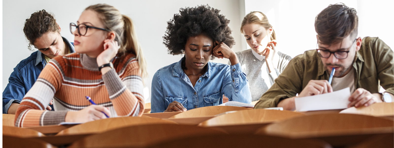 Five students sitting at desks in a classroom, avoiding the flu, appear focused as they write or read, with notebooks and papers in front of them. The classroom is well-lit with natural light.