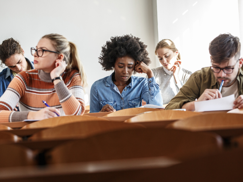 How to avoid the flu. A group of five students sitting in a classroom, focused on writing or taking a test at their desks. Sunlight streams in from a window, creating a bright, studious atmosphere.